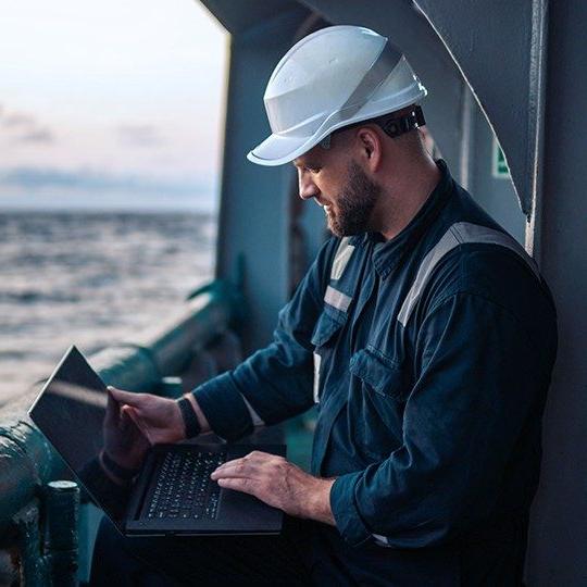 Man in a hard hat on a vessle at sea using maritime internet on a laptop.